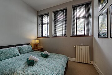 A modern bedroom with a bed featuring green patterned bedding, a nightstand with a lamp, three windows with blinds, a radiator, and wall art.
