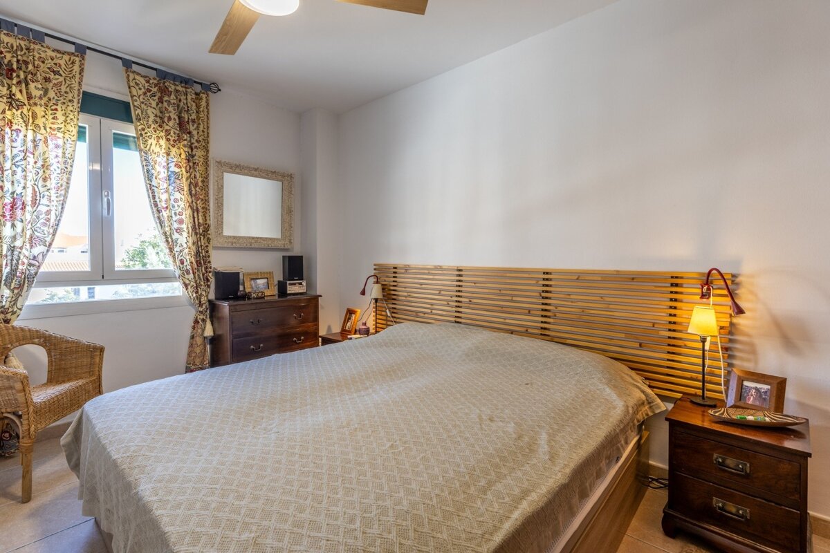 A cozy bedroom with a bed, slatted wooden headboard, patterned curtains, wooden dresser, and nightstands. A ceiling fan is visible above.