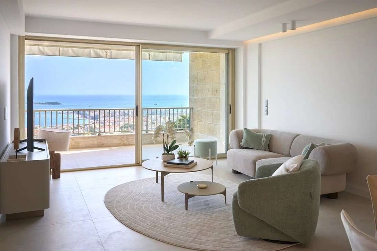 A chic, modern living room with neutral-toned furniture and a round rug, featuring large sliding doors that open to a balcony with a panoramic ocean and city view.