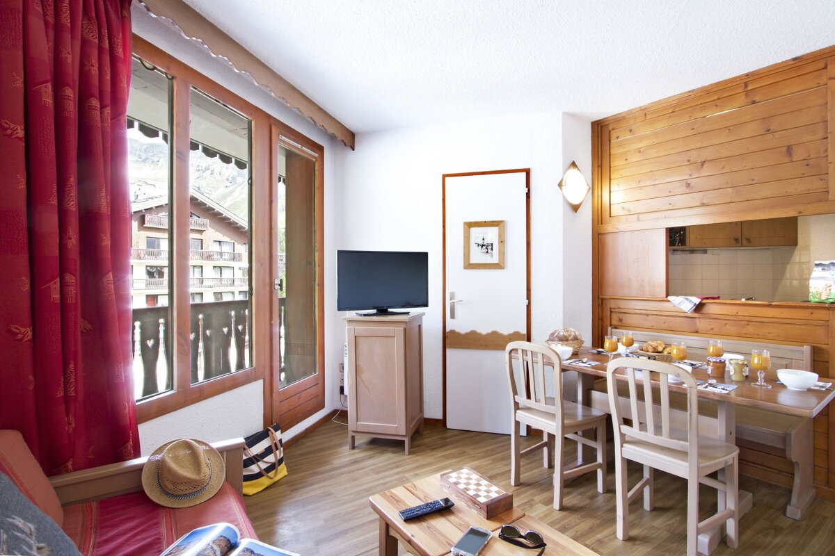 A cozy, wood-accented living/dining room with a breakfast spread on the table, red curtains, and a balcony offering a view of chalets and mountains.