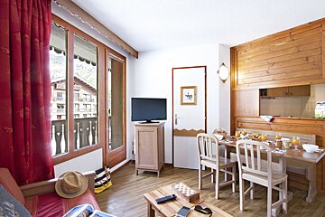 A cozy, wood-accented living/dining room with a breakfast spread on the table, red curtains, and a balcony offering a view of chalets and mountains.