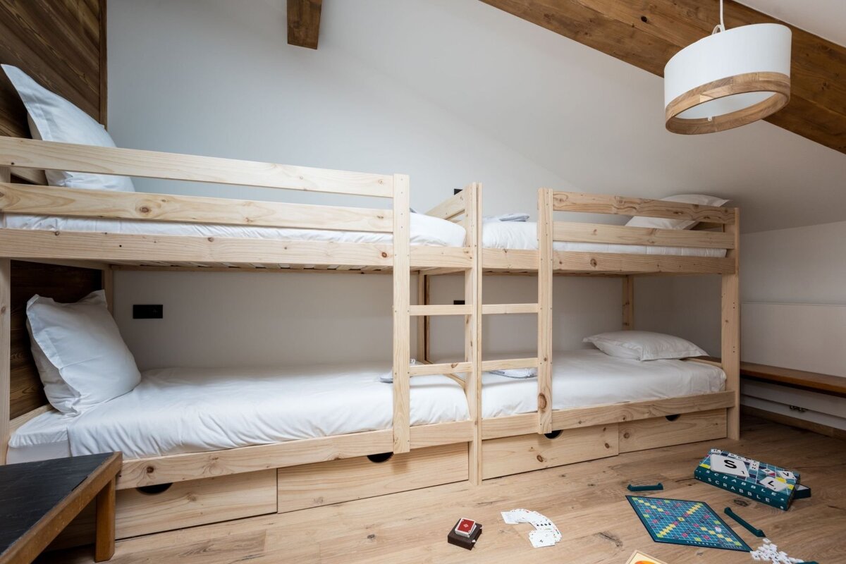 A rustic room with two wooden bunk beds and white bedding. Exposed wooden beams adorn the ceiling, and board games are scattered on the floor.