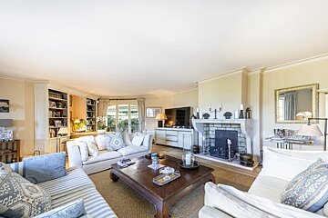 A cozy, sunlit living room featuring cream and blue sofas, a large wooden coffee table, fireplace, TV, and built-in bookshelves.