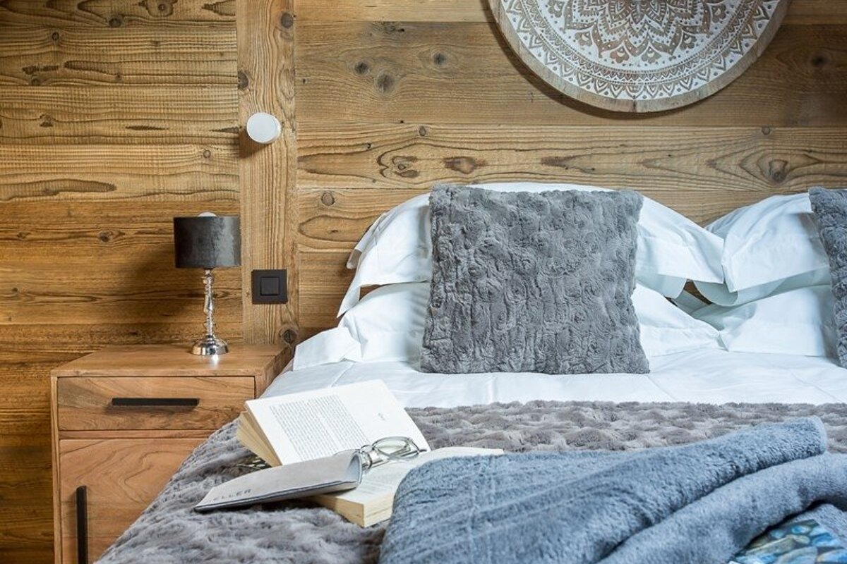 Cozy chalet-style bedroom with wood walls, soft grey bedding, an open book, and a wooden nightstand with a lamp.