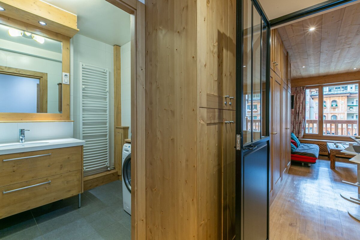 A modern apartment interior with light wood, featuring a bathroom with a washing machine on the left and a living room with large windows on the right.