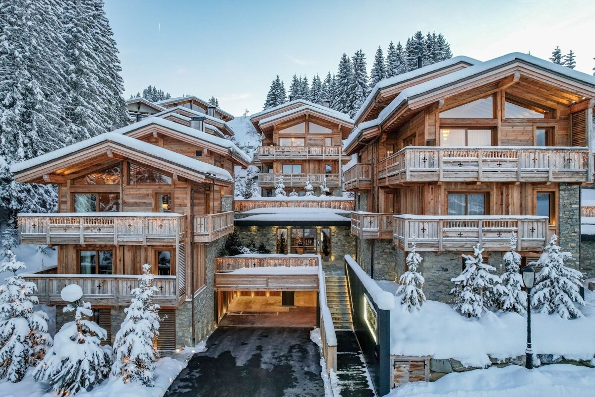 Luxury timber chalets covered in snow, nestled within a pine forest, with illuminated balconies and an impressive entrance in a winter landscape.