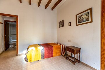 A bedroom with a single bed, colorful blanket, nightstand, paintings, and exposed wooden beams. An open door reveals a bathroom with a toilet.