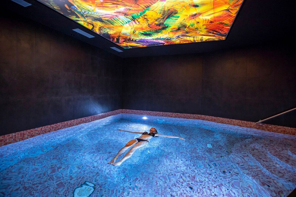 A woman floats serenely in a dimly lit pool under a vibrant, colorful abstract ceiling light display, creating a relaxing spa ambiance.