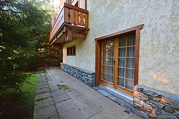 A stucco building with a stone base, wooden balcony, and glass doors, alongside a paved path and lush green trees, bathed in sunlight.