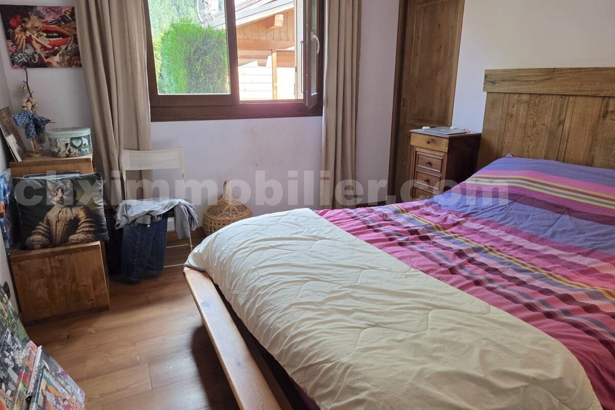 A cozy bedroom features a wooden bed with colorful striped bedding, a wooden headboard, and a nightstand. A window with curtains offers a view outside.