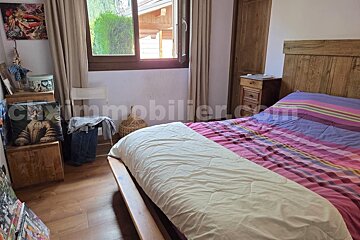 A cozy bedroom features a wooden bed with colorful striped bedding, a wooden headboard, and a nightstand. A window with curtains offers a view outside.