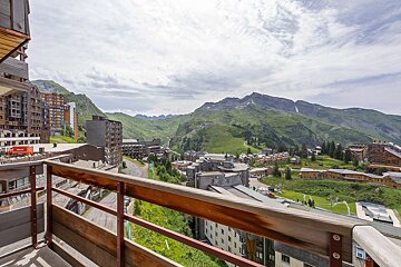 Balcony view of a mountain resort. Buildings dot green valleys, flanked by vast mountains with snow patches under a cloudy sky.