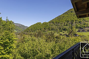 A sunny balcony view overlooks a lush green mountainous valley with dense forests, a clear blue sky, and a small village nestled in the distance.
