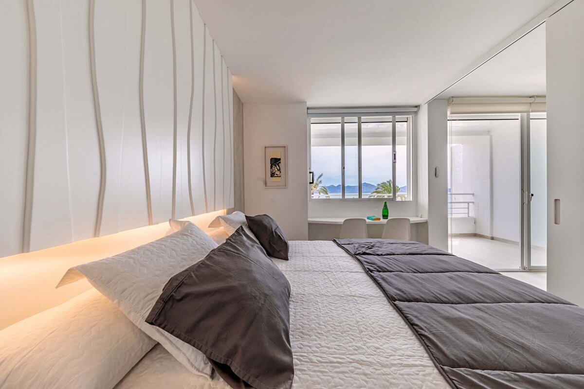 A modern, light-filled bedroom with a wavy textured wall, backlit headboard, and a large window offering sea and mountain views.
