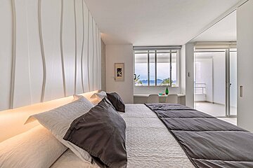A modern, light-filled bedroom with a wavy textured wall, backlit headboard, and a large window offering sea and mountain views.