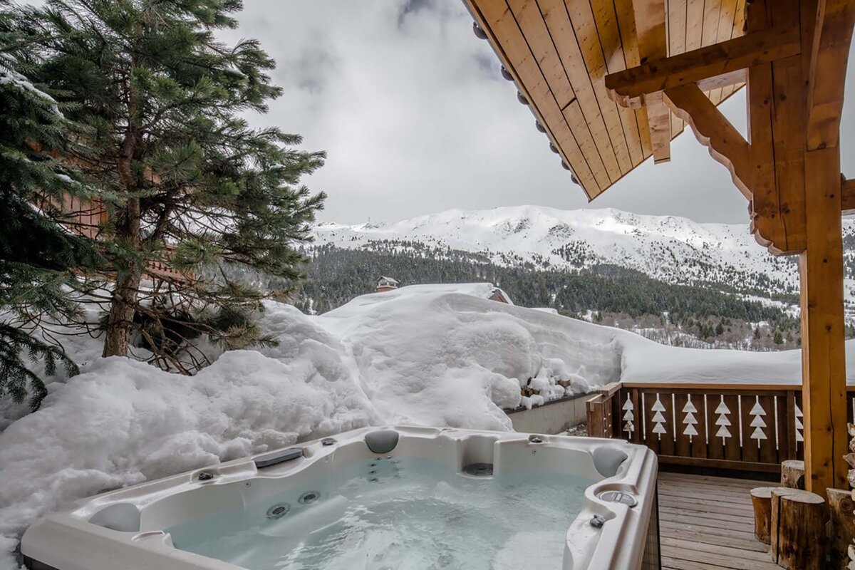 A hot tub on a snowy wooden deck with a pine tree, overlooking snow-covered mountains and a valley under a cloudy winter sky.