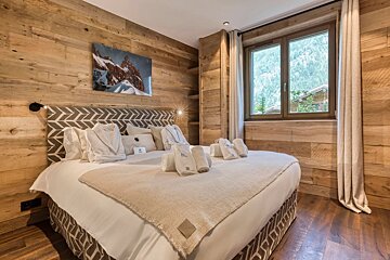 Cozy chalet bedroom featuring warm wood-paneled walls, a large bed with white linens, and a window with mountain views.