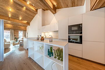 Modern kitchen with white cabinets, a vaulted wooden ceiling, and a storage island, looking out towards a bright living area.