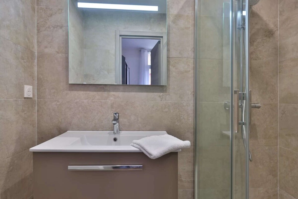 Modern bathroom features a sink with a white basin and brown vanity, a mirror with a light strip, and a glass-enclosed shower. Tiled walls in neutral tones.