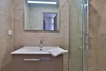 Modern bathroom features a sink with a white basin and brown vanity, a mirror with a light strip, and a glass-enclosed shower. Tiled walls in neutral tones.