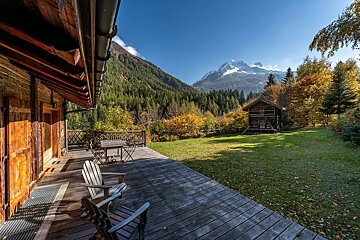 A rustic wooden chalet with a large deck offers a stunning view of vibrant autumn forests and majestic snow-capped mountains under a clear sky.