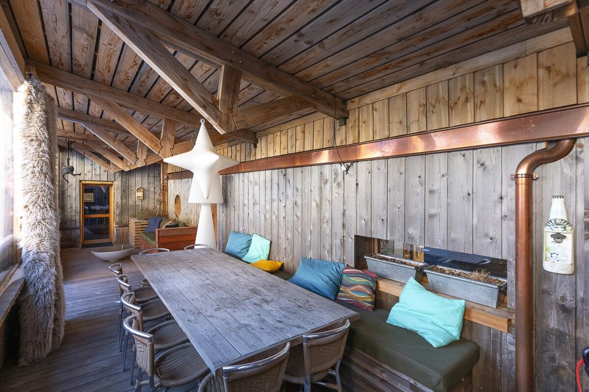 Rustic wooden covered patio featuring a long dining table, seating with colorful pillows, and exposed beams overhead.
