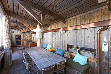 Rustic wooden covered patio featuring a long dining table, seating with colorful pillows, and exposed beams overhead.