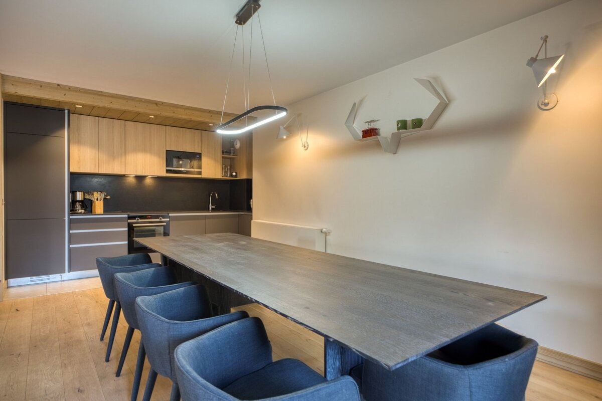 Modern dining area featuring a long dark wood table, blue armchairs, and a sleek kitchen in the background.