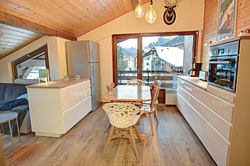 Modern chalet dining area and kitchen, featuring a wooden table and large glass doors looking out onto a snowy mountain view.