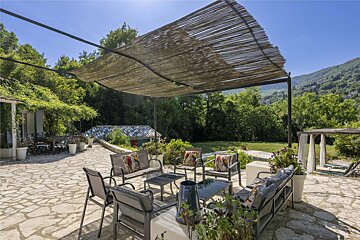 A sunny stone patio with lounge furniture under a bamboo pergola, surrounded by lush green trees and distant hills, offering a serene outdoor space.