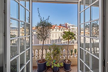 View of a city balcony with potted plants through open French doors under a clear blue sky.
