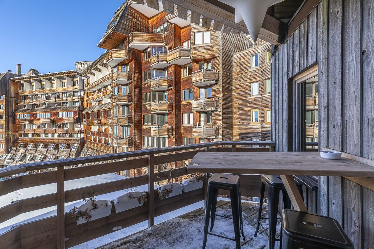 A snow-dusted wooden balcony with a high table and stools offers views of large timber ski resort buildings.