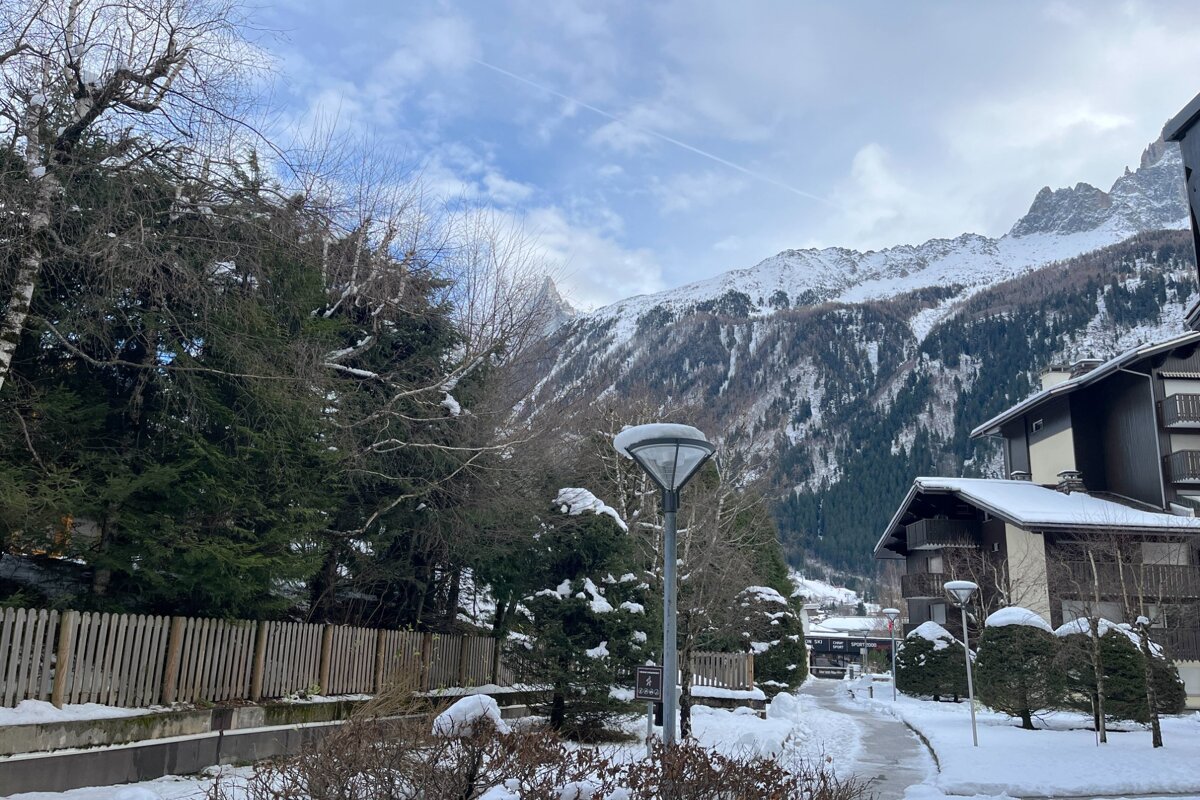 A cold winter view showing snow-covered mountains, evergreen trees, chalets, and a snowy path under a cloudy sky.