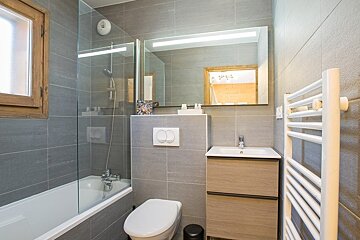 Modern grey-tiled bathroom featuring a glass-enclosed bathtub, toilet, wooden vanity with sink, and a large illuminated mirror.