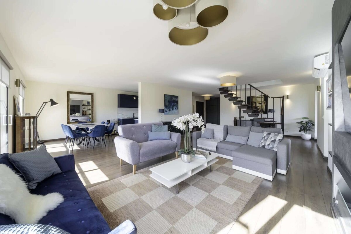 A bright, open-plan living room with blue and grey sofas, a dining area, and a kitchen. Modern decor and a staircase complete the stylish space.