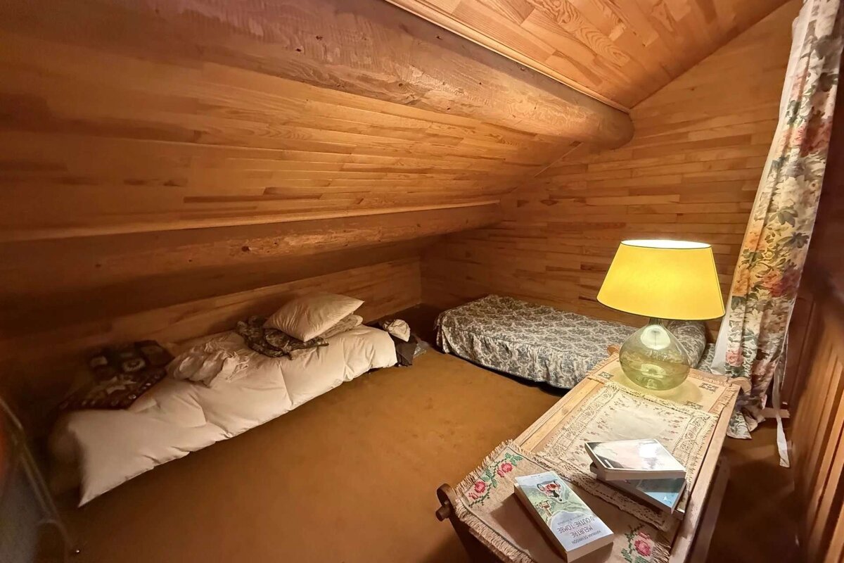 A cozy, wood-paneled attic with a slanted ceiling, two floor beds, a lamp on a small table with books, and a floral curtain.