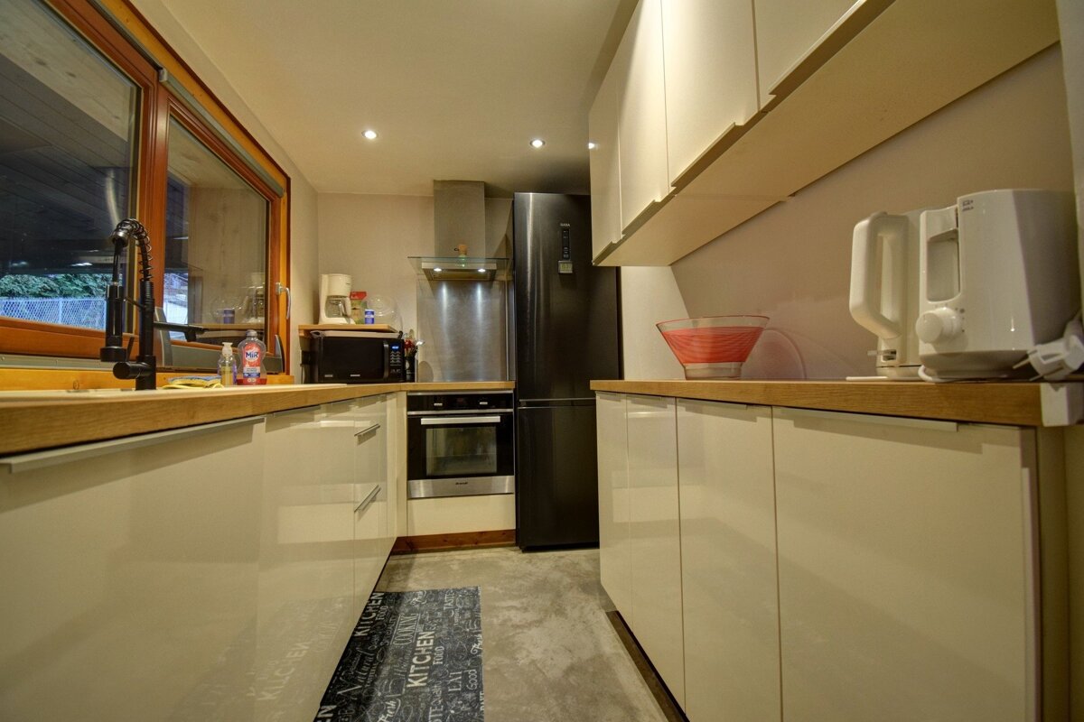 Modern galley kitchen featuring glossy off-white cabinets, light wood countertops, and a tall black refrigerator at the far end.