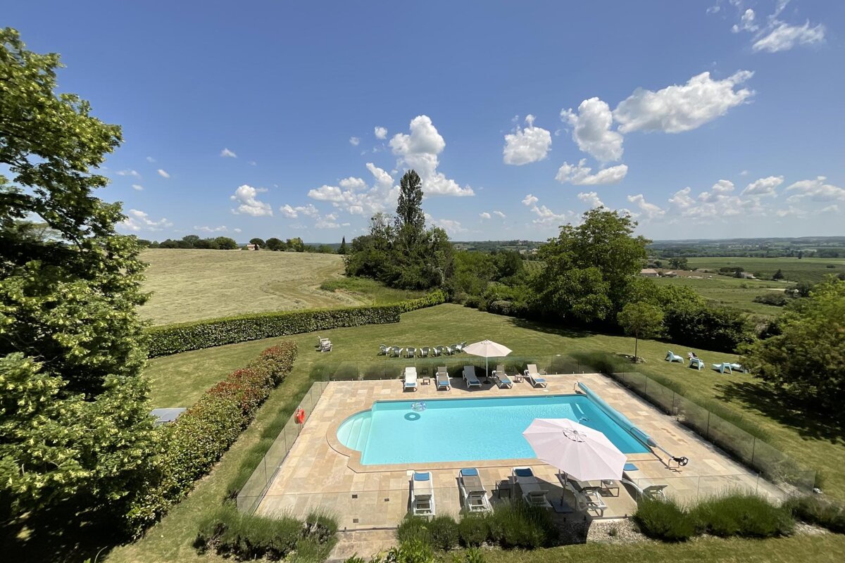High-angle view of a swimming pool with lounge chairs on a sunny day, surrounded by lush green lawns, fields, and trees under a blue, cloudy sky.