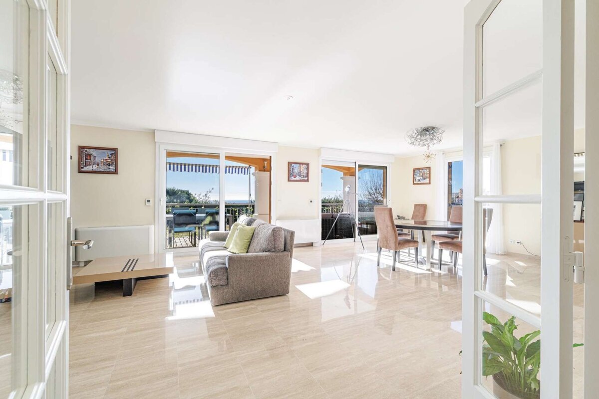 A bright, spacious living and dining room with a light tile floor, sofa, dining table, and large sliding doors opening to a balcony with a scenic view.