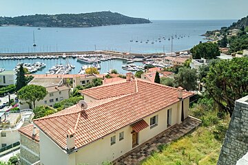 A scenic view of a Mediterranean coastal town with terracotta-roofed houses overlooking a harbor filled with boats and the blue sea.