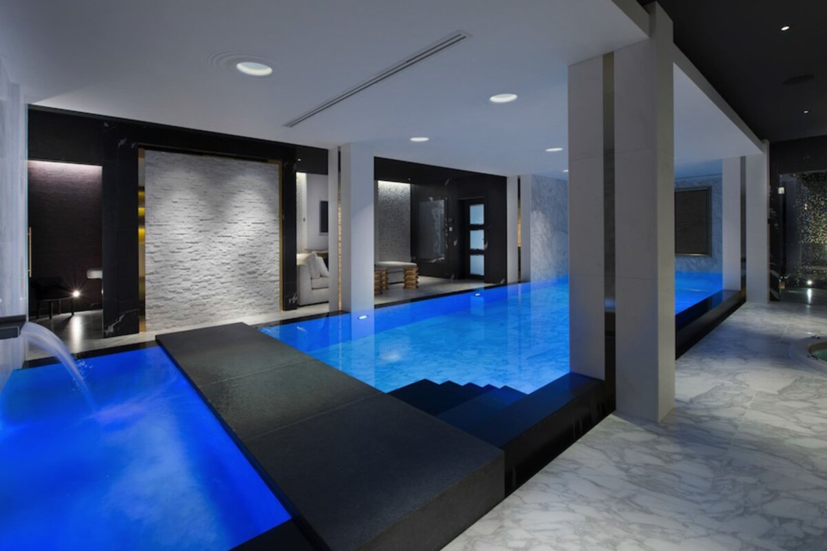 A sleek indoor pool area featuring luminous blue water, dark and light marble, a modern footbridge, and a water spout, creating a luxurious spa ambiance.