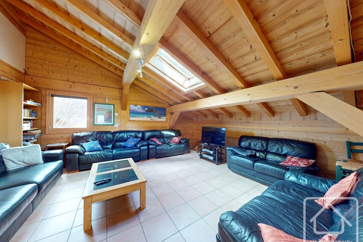 A cozy wooden attic living room with black leather sofas, a coffee table, tiled floor, exposed beams, a skylight, and a window.