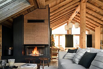 Cozy modern cabin living room featuring a burning fireplace, exposed wood-beamed ceilings, and a comfortable grey sofa.