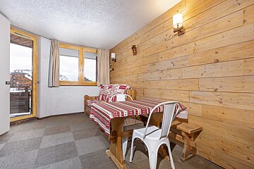 Chalet living area with wood paneling, dining table, sofa, and a snowy mountain view from the window and balcony.