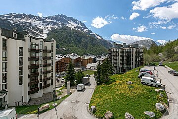 Modern buildings & traditional chalets in an alpine town, surrounded by snowy mountains, green hills, and a bright blue sky.