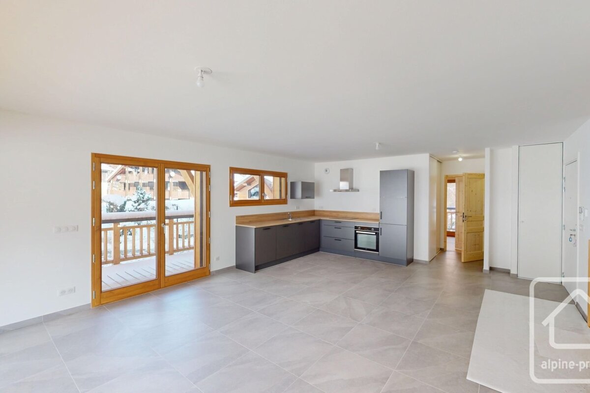 An empty, modern apartment with tiled floors, a grey kitchen, and sliding wooden glass doors leading to a balcony.