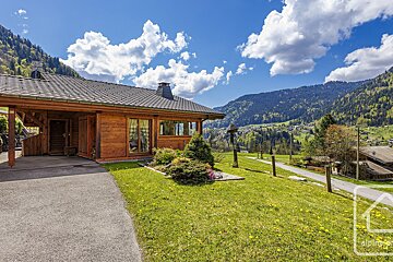 A wooden house sits on a lush green hillside with mountains in the background