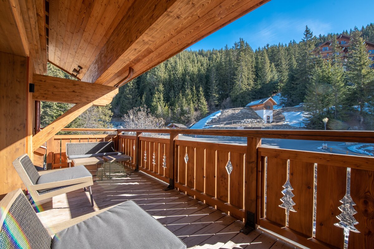 Wooden chalet balcony with comfortable chairs overlooking a dense pine forest and snowy mountains on a sunny day.