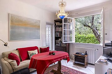 A cozy living/dining room features a red-covered table, sofa, chandelier, and an open window revealing a lush green garden view.