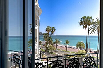 Balcony view of a vibrant turquoise sea, palm trees, and a promenade next to a grand white building under a clear blue sky.